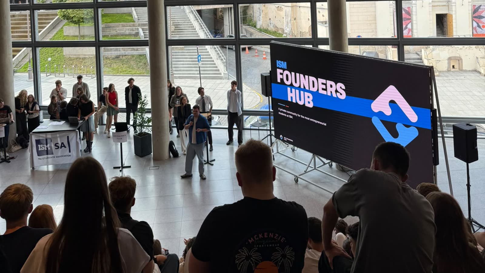 Studentai ISM atriume per Club Fair, priešais ISM Founders Hub ekraną